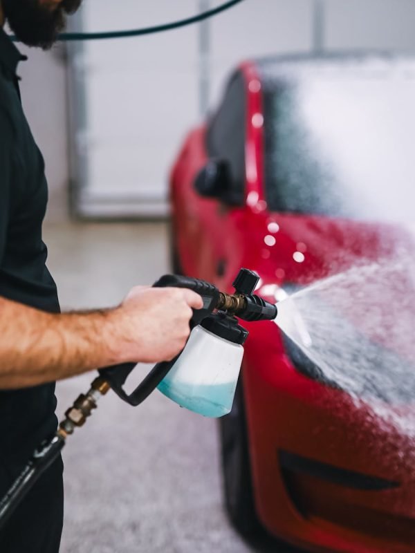 A man uses a foam sprayer to clean a red car indoors, showcasing car detailing.